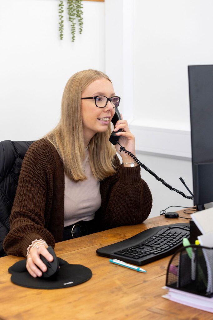brown cardigan, plant, white wall, desk, recruiter, on the phone, keyboard, mouse, screen, pen, outsourced recruitment, blonde hair, glasses, woman, at work, wooden desk, recruitment, telephone interview 