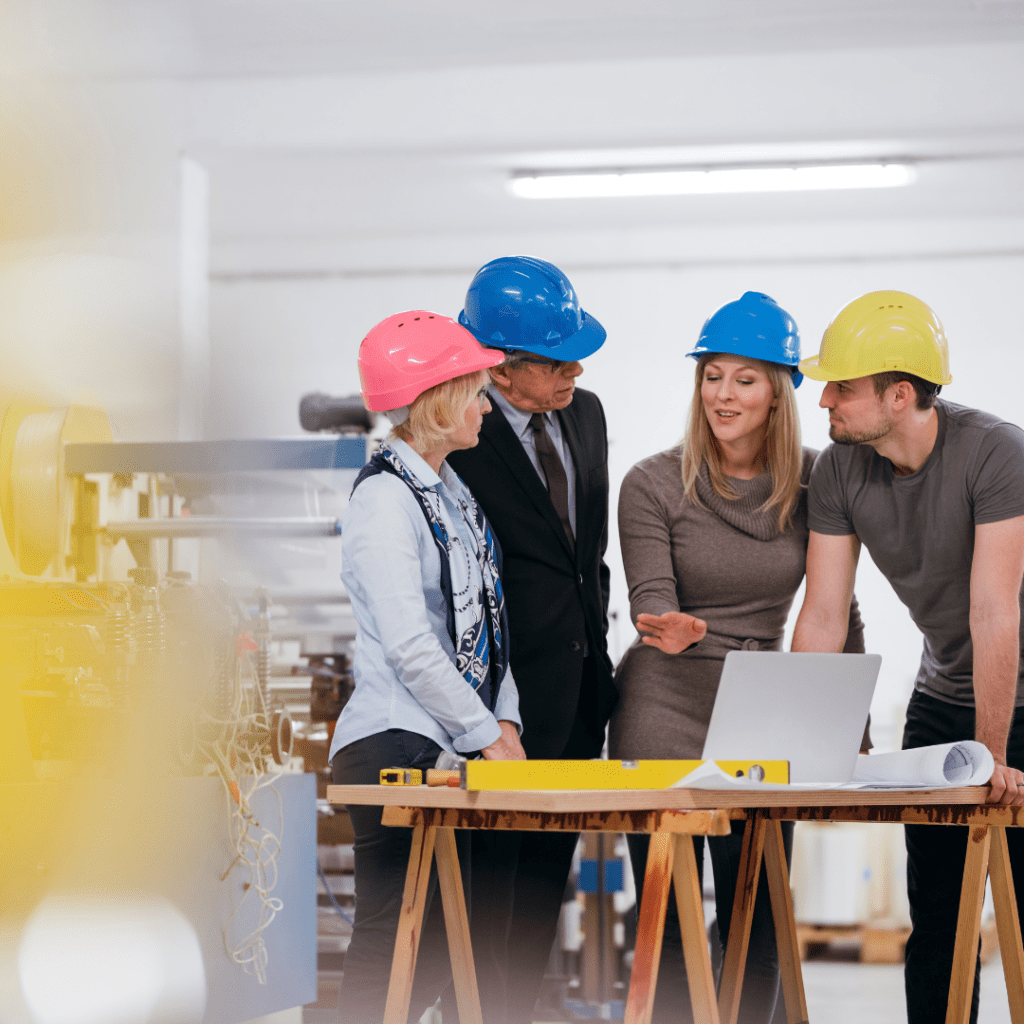 team, builders, construction, hard hats, yellow hard hat, blue hard hat, pink hard hat, table, plans, laptop, workplace, maintenance, 4 people, women, men, leadership, management, problem solving 